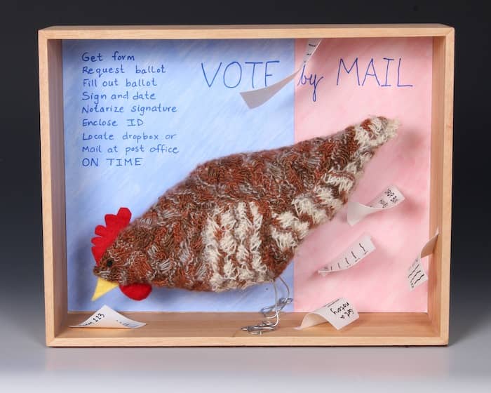  brown knitted chicken against backdrop of mail-in-voting instructions with papers flying in air 9 x 12 x 4 inches ©2022 Eve Jacobs-Carnahan
