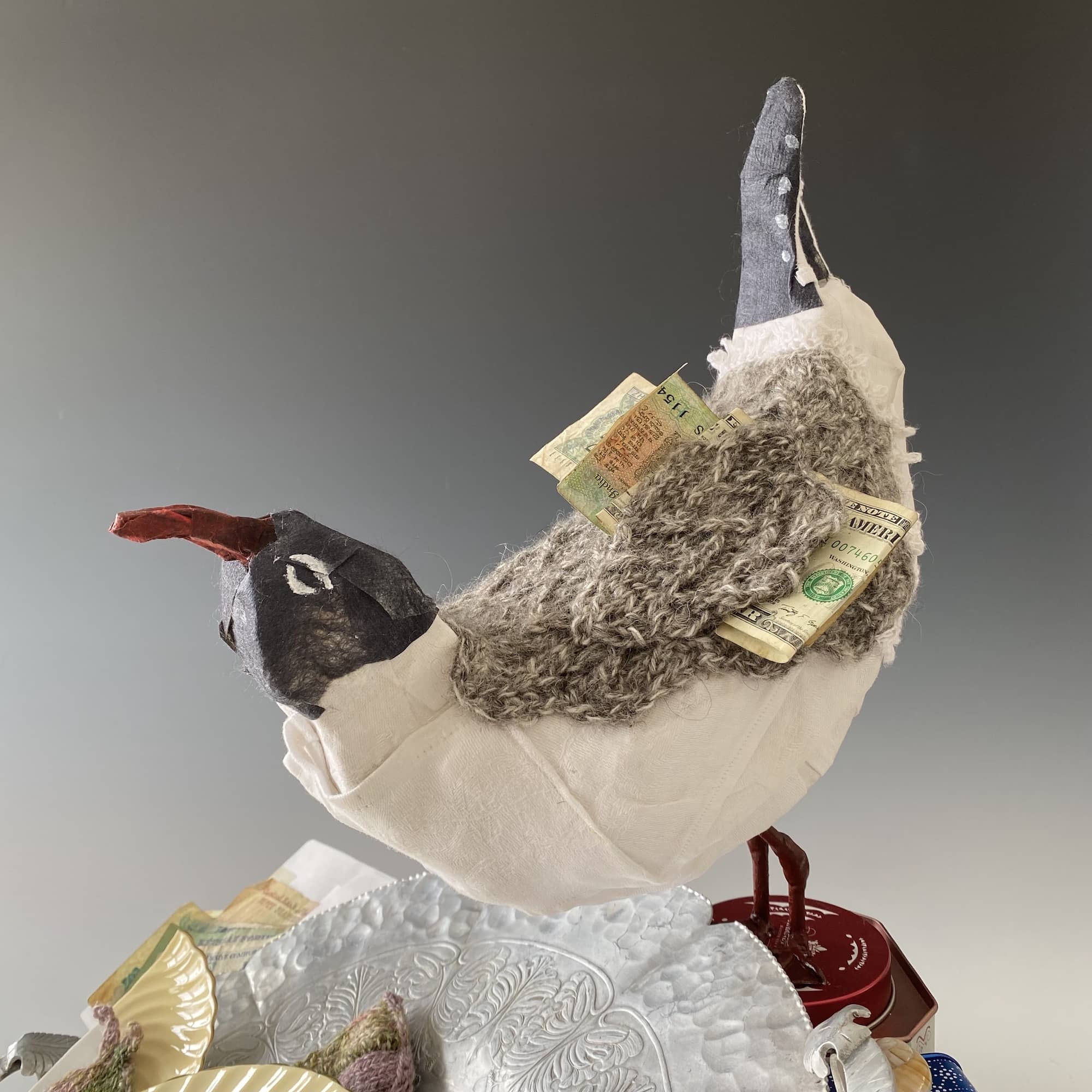 mixed media sculpture of laughing gull with black silk head, white linen breast, knitted wool body, with currency tucked under wing