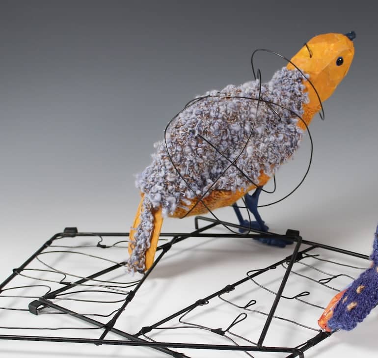 fanciful bird sculpture with curly boucle yarn back, yellow orange body, surrounded by haphazard wire rings standing on edge of  broken metal cage