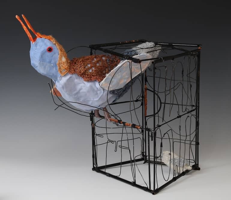 fanciful sculpture of blue, orange, grey bird breaking through wires of black metal enclosure