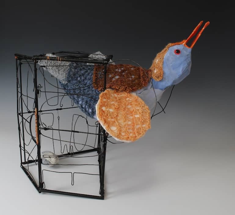 sculpture of fanciful gray, blue, orange bird balanced between inside and outside of cage with tiny gray bird remaining at bottom corner of cage