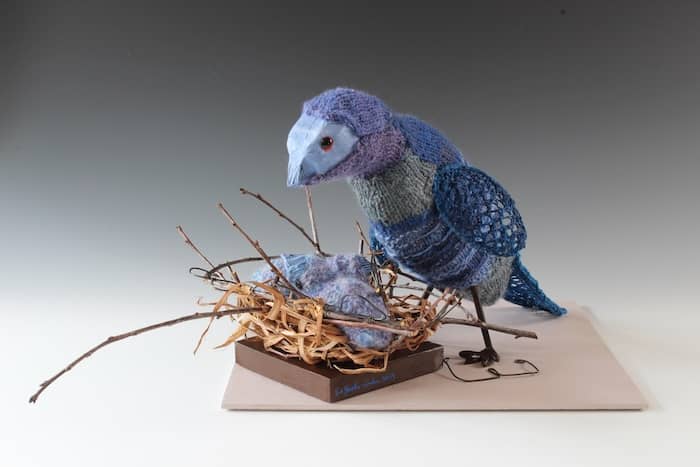 blue knitted bird standing over chicks in nest of twigs, reeds, and wire letters